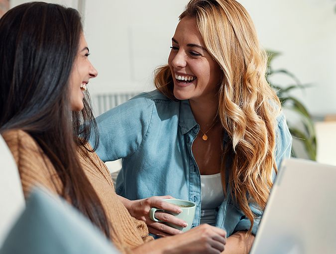 Twee vrouwen zitten samen lachend op een bank. De een houdt een mok vast, terwijl de ander een laptop op schoot heeft.