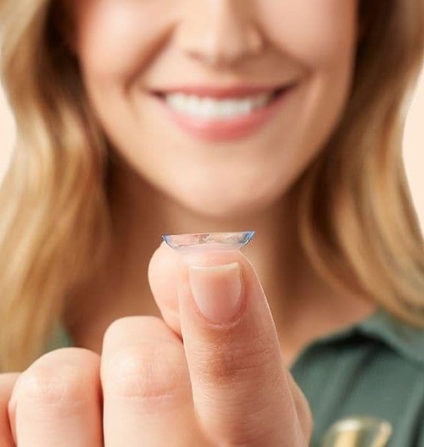 Plan rapproché sur une femme regardant une lentille posée sur son doigt avec un sourire aux lèvres. On ne voit pas ses yeux.
